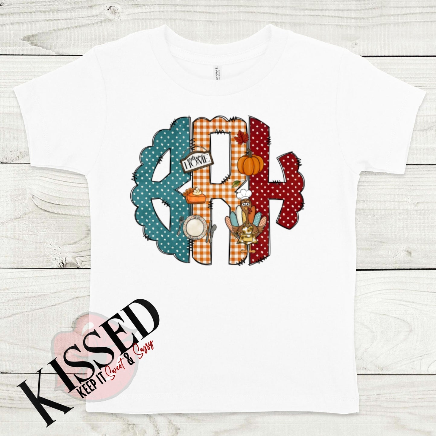 A stylish fall staple, this white Fall Monogram Shirt from Keep It Sweet & Sassy Designs features a large "RH" in teal, red, and orange checkered patterns adorned with a small pumpkin, leaves, and a cup of cocoa. Laid flat on a wooden surface with the "KISSED" logo at the bottom left, it's made from high-quality polyester.