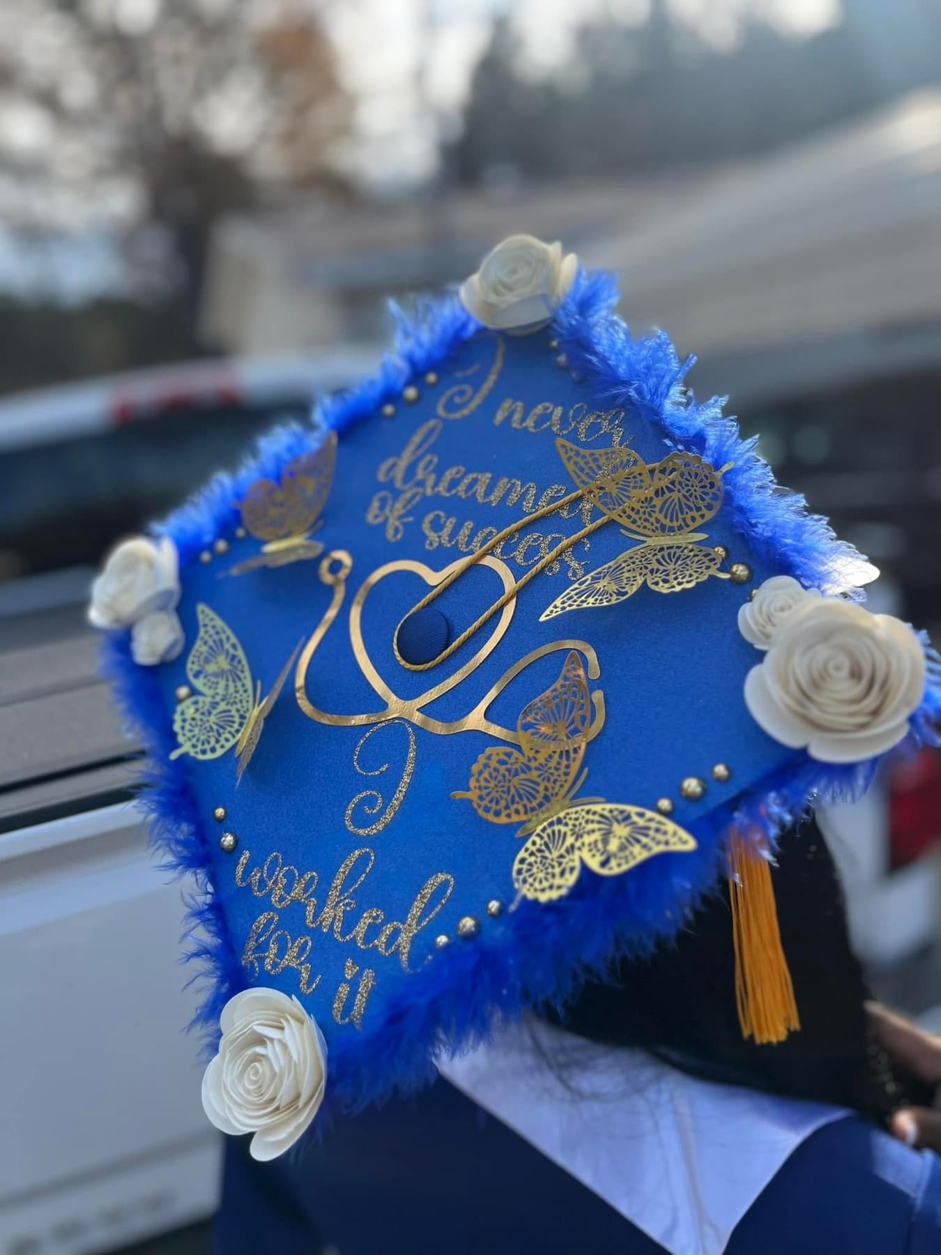 The "Graduation Cap" by Keep It Sweet & Sassy Designs is a personalized accessory adorned with white roses and gold embellishments, including butterflies and leaves. It features the inspiring quote: "I never dreamed of success, I worked for it." Blue feathers line the edges, and a yellow tassel hangs on the side to showcase graduation pride.