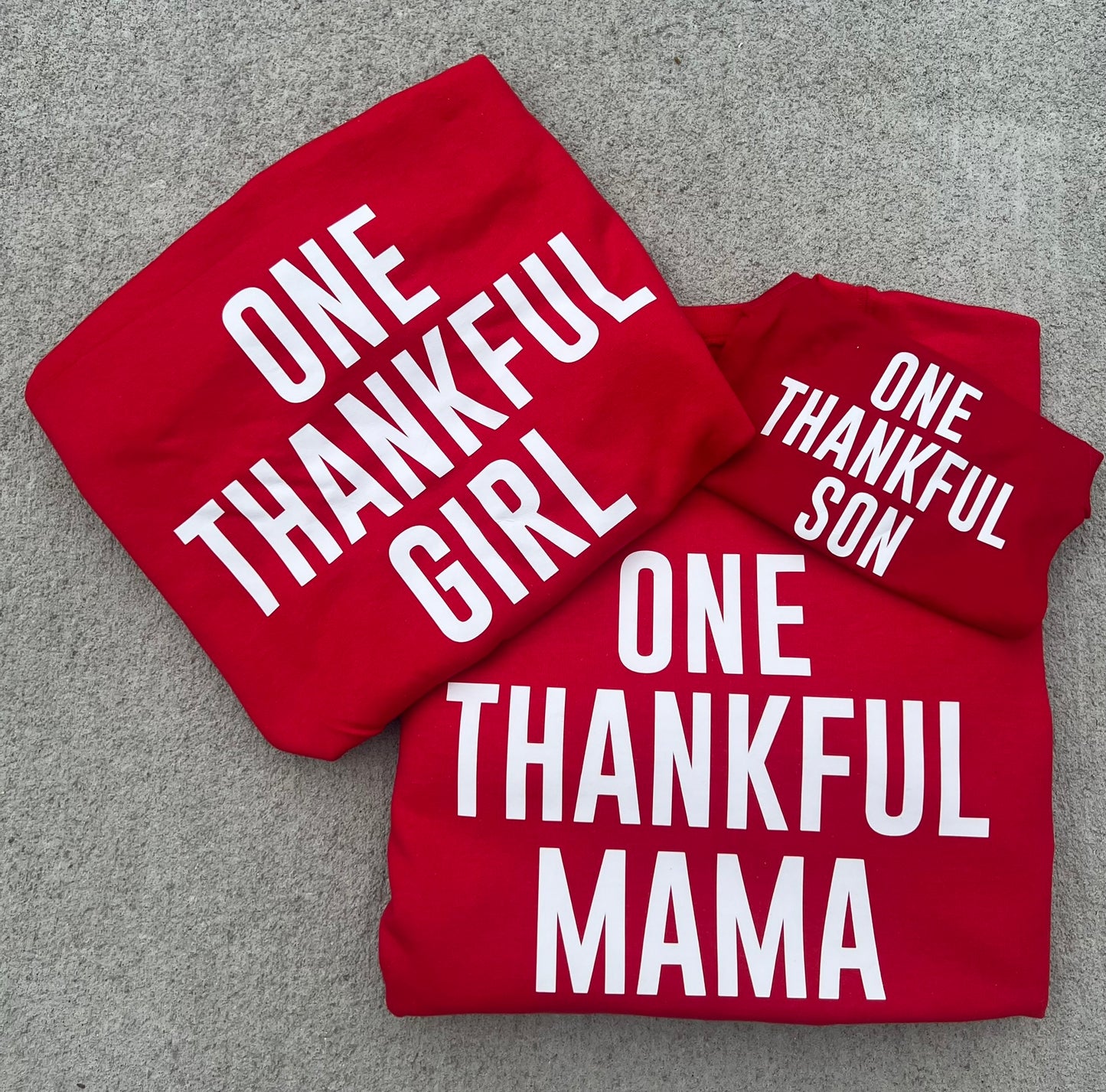 Three stylish, custom sweaters from Keep It Sweet & Sassy Designs are displayed on a light gray surface. Each red sweater features white text reading "ONE THANKFUL GIRL" on the left, "ONE THANKFUL SON" on the right, and "ONE THANKFUL MAMA" at the bottom. Perfect for fall and winter, these pieces showcase your gratitude in style.