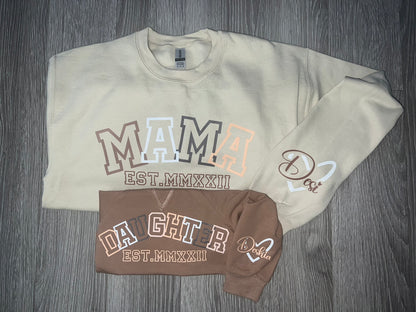 Two stylish "Keep It Sweet & Sassy Designs" Custom Sweaters are displayed on a wooden surface. The larger, beige sweater reads "MAMA EST. MMXXII" with a heart and the name "Dori," while the smaller, brown sweatshirt reads "DAUGHTER EST. MMXXII" with a heart and the name "Dahlia." Perfect for fall and winter coziness.