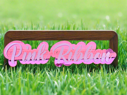 A sign that reads "Pink Ribbon" in stylized pink letters, crafted by Keep It Sweet & Sassy Designs, is mounted on a wooden frame. The Acrylic Name Sign adds a professional touch, set against a backdrop of lush green grass.