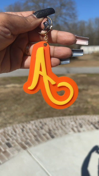 A hand with long, dark nails holds a Keep It Sweet & Sassy Designs Acrylic Letter Keychain, featuring a stylized, cursive letter 'A' in yellow and orange. The background shows an outdoor scene with grass, a pathway, and trees in the distance, hinting at an adventurous side.