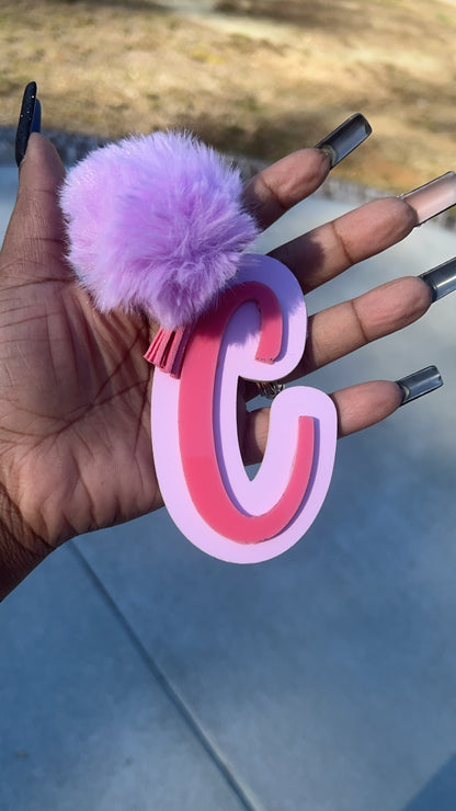 A hand with long, dark nails holds a Keep It Sweet & Sassy Designs Acrylic Letter Keychain, featuring a stylized, cursive letter 'A' in yellow and orange. The background shows an outdoor scene with grass, a pathway, and trees in the distance, hinting at an adventurous side.