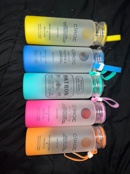 Five vibrant drink bottles are laid out horizontally on a black surface. Each bottle, resembling the Glass Alcohol Tumbler from Keep It Sweet & Sassy Designs, features a brand name and comes in striking shades of yellow, gray, blue, pink, and orange. The lids have matching wrist straps that enhance their sleek design.