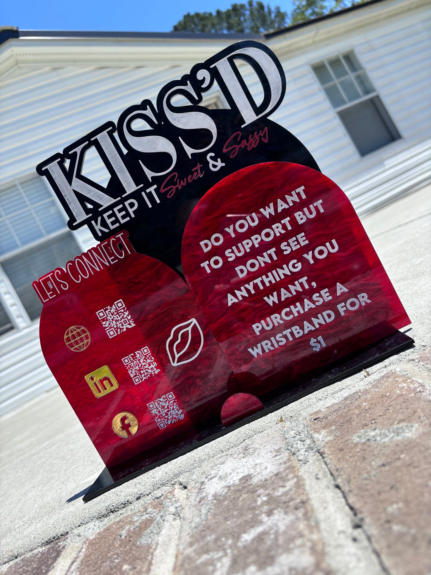 A stand-up sign outdoors advertises "Keep It Sweet & Sassy Designs." It encourages support through wristband purchases and includes icons for social media platforms like Facebook, Instagram, and LinkedIn, along with two QR codes. The background features a house and a brick surface with an Acrylic Payment Sign.