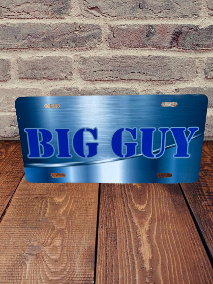 Displayed against a backdrop of a brick wall and wooden floor is the Car Tag by Keep It Sweet & Sassy Designs, featuring a personalized metallic blue finish. The plate proudly showcases the words "BIG GUY" in large, bold blue letters and includes four mounting holes at the top and bottom for easy installation, adding an elegant touch to any vehicle.