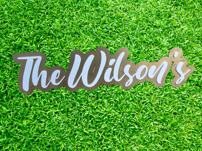 A sign that reads "Pink Ribbon" in stylized pink letters, crafted by Keep It Sweet & Sassy Designs, is mounted on a wooden frame. The Acrylic Name Sign adds a professional touch, set against a backdrop of lush green grass.