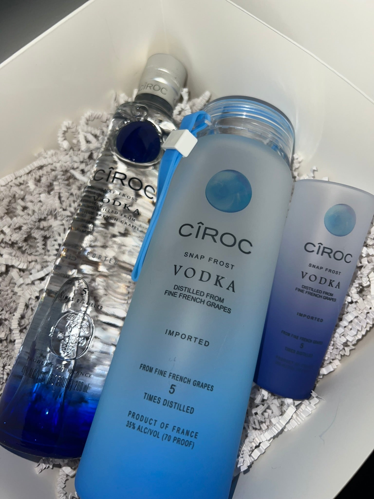 A display featuring a trio of CÎROC Snap Frost Vodka bottles in a pristine white gift box lined with shredded paper. The sleek design showcases tall, slender bottles; one transparent with a blue gradient and two with subtle blue hints. Each bottle features consistent branding and is accompanied by an elegant Glass Alcohol Tumbler from Keep It Sweet & Sassy Designs, perfect for the ideal pour.