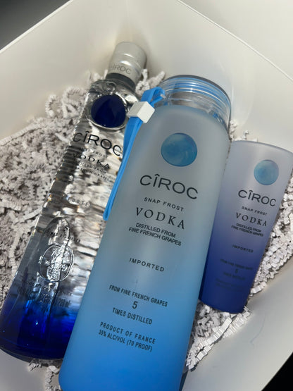 A display featuring a trio of CÎROC Snap Frost Vodka bottles in a pristine white gift box lined with shredded paper. The sleek design showcases tall, slender bottles; one transparent with a blue gradient and two with subtle blue hints. Each bottle features consistent branding and is accompanied by an elegant Glass Alcohol Tumbler from Keep It Sweet & Sassy Designs, perfect for the ideal pour.